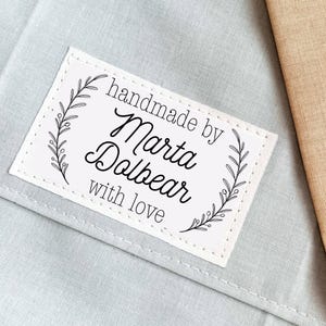 Peut inclure: Une étiquette rectangulaire blanche avec le texte "handmade by Marta Dolbear with love" en écriture noire, encadrée d'un motif floral délicat. L'étiquette est cousue sur un tissu bleu clair.