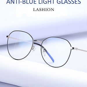 Puede incluir: Gafas redondas de bloqueo de luz azul con montura plateada y puente nasal azul. Las gafas est&aacute;n sobre una superficie blanca. ANTI-BLUE LIGHT GLASSES LASHION
