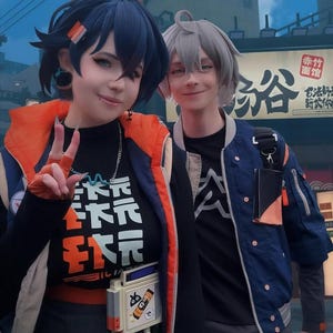 May include: Two people in cosplay outfits pose for a photo. One wears a black shirt with orange and white text, an orange and blue jacket, and a silver chain. The other wears a blue jacket and a black shirt with a white graphic.