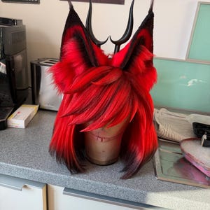 Op de afbeelding: Een rood-zwarte cosplaypruik met bevestigde zwarte hoorns en oren. De pruik heeft lang, gelaagd rood haar met zwarte punten. De oren zijn zwart met rood haar aan de binnenkant. De hoorns zijn gebogen en zwart.