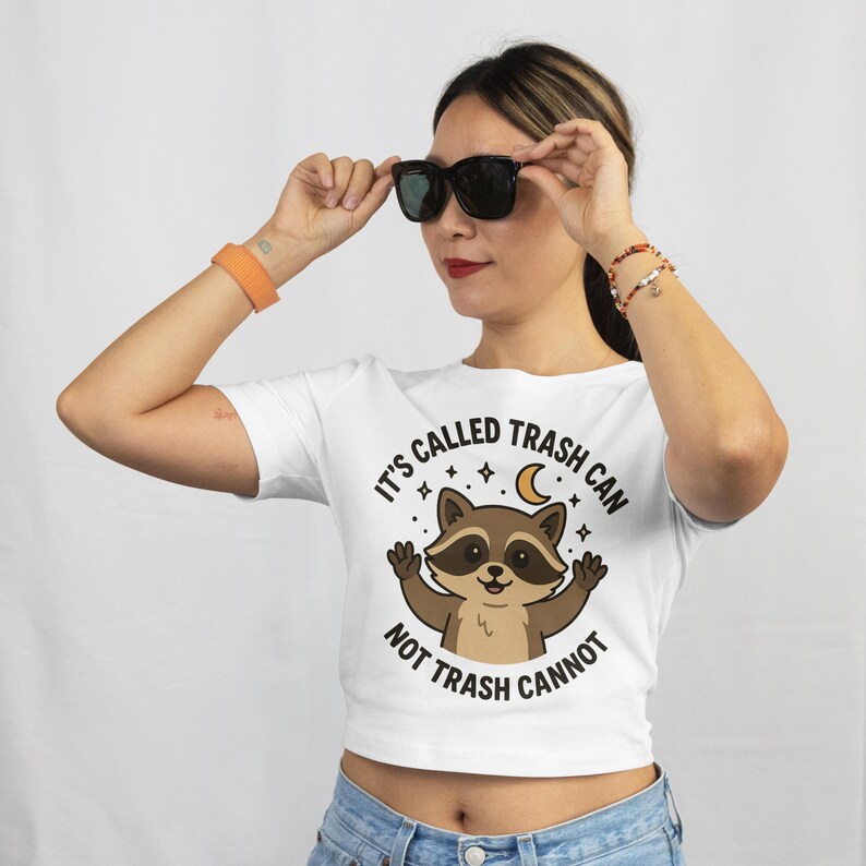 May include: White crop top featuring a cartoon raccoon with the text "IT'S CALLED TRASH CAN NOT TRASH CANNOT". The raccoon is brown and tan with a smiling face. The shirt is worn with blue jeans and black sunglasses.
