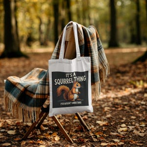 Op de afbeelding: Witte draagtas met een afbeelding van een eekhoorn en de tekst "It's a Squirrel Thing (You Wouldn't Understand)". De tas hangt aan een houten stoel met een geruite deken. De achtergrond is een vage bossc&egrave;ne.