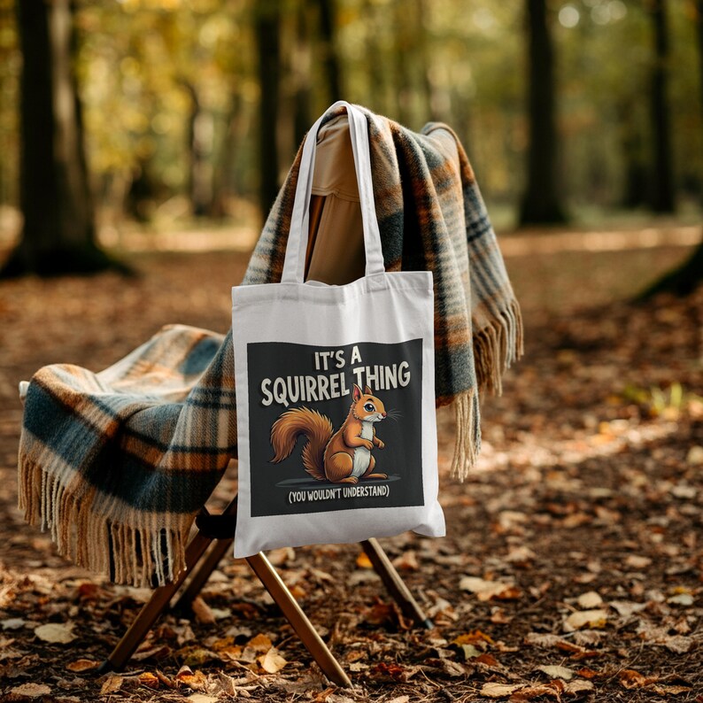 Op de afbeelding: Witte draagtas met een afbeelding van een eekhoorn en de tekst "It's a Squirrel Thing (You Wouldn't Understand)". De tas hangt aan een houten stoel met een geruite deken. De achtergrond is een vage bossc&egrave;ne.