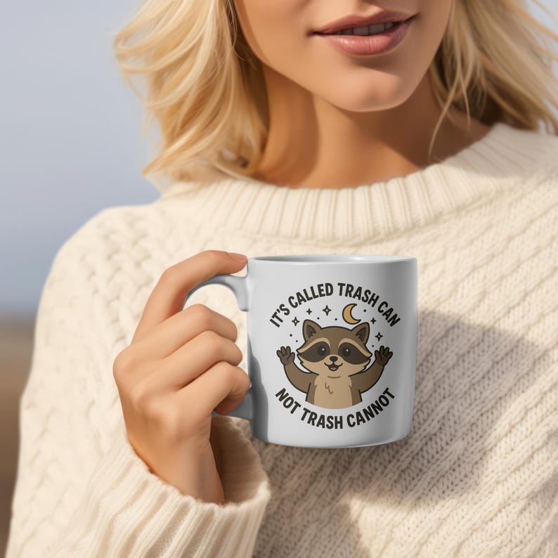 May include: White ceramic mug featuring a cartoon raccoon with outstretched arms, a crescent moon, and stars. The mug has the text "IT'S CALLED TRASH CAN NOT TRASH CANNOT" in a circular design.