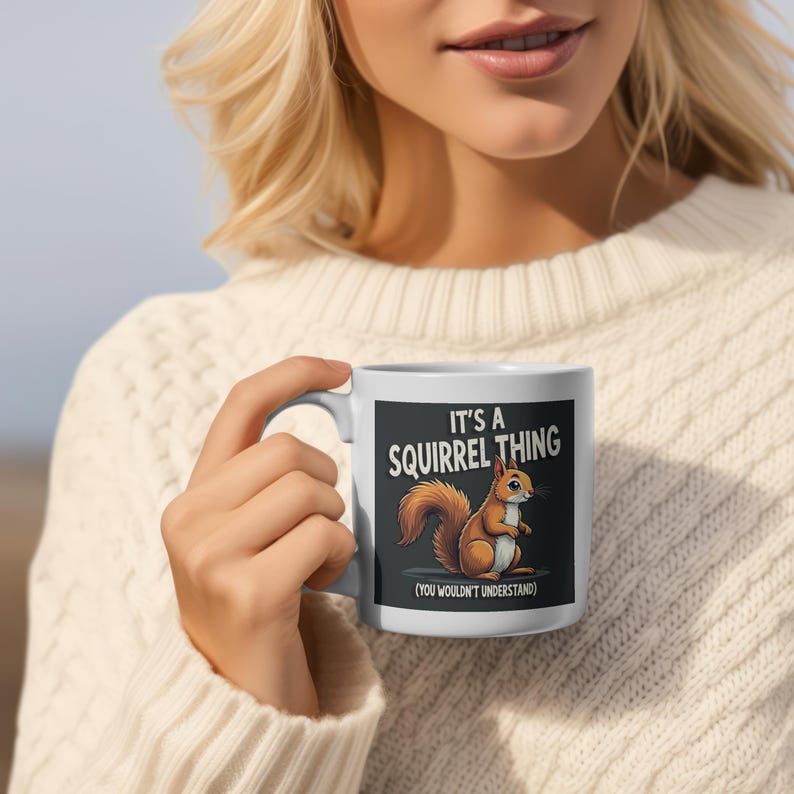 Op de afbeelding: Witte keramische mok met een cartoon eekhoorn ontwerp en de tekst "IT'S A SQUIRREL THING (YOU WOULDN'T UNDERSTAND)". De eekhoorn is bruin en oranje. De mok heeft een wit handvat.
