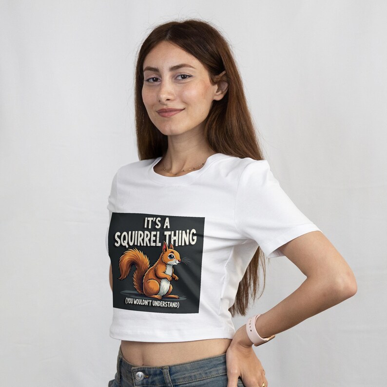 Op de afbeelding: Witte crop top met een cartoon eekhoorn afbeelding en de tekst "IT'S A SQUIRREL THING (YOU WOULDN'T UNDERSTAND)". De eekhoorn is bruin en oranje, tegen een zwarte achtergrond. Het shirt wordt door een persoon gedragen.