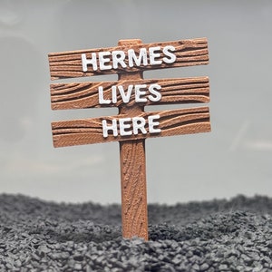 Puede incluir: Un letrero de madera con tres líneas de texto que dice "HERMES LIVES HERE" en letras blancas. El letrero está de pie en un lecho de grava negra.