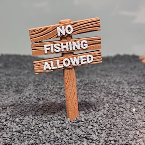 Peut inclure: Un panneau en bois avec le texte "NO FISHING ALLOWED" en lettres blanches. Le panneau est posé sur un lit de petits cailloux noirs.