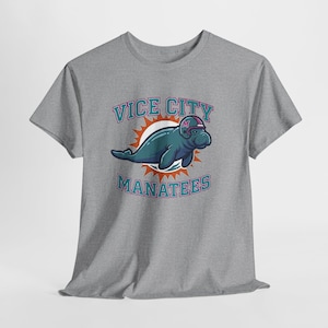 Op de afbeelding: Een grijs gemêleerd t-shirt met korte mouwen en een afbeelding van een talingkleurige zeekoe met een footballhelm. Het ontwerp bevat een oranje zonnestraal-achtergrond en de tekst VICE CITY MANATEES in talingkleurige en paarse letters op de voorzijde.