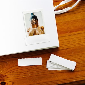 Peut inclure: Un album photo blanc avec une photo Polaroid d'une personne se couvrant le visage avec les mains. La photo est sur une surface en bois avec deux embellissements en papier festonnés blancs.