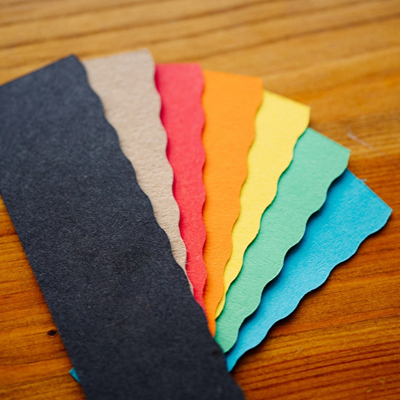 May include: A fan-shaped arrangement of seven different colored paper strips. The strips are black, brown, red, orange, yellow, green, and blue. The strips are all the same size and shape, and they are arranged in a way that creates a rainbow effect.