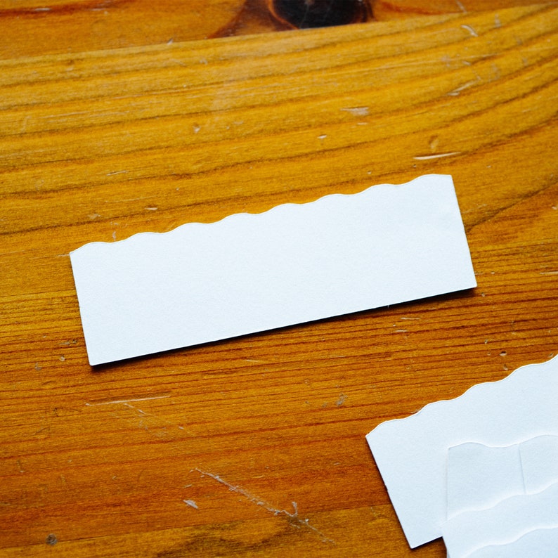 May include: Two white scalloped edge paper cards on a wooden surface. The cards are blank.