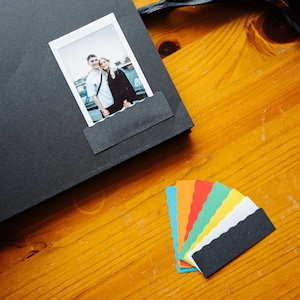 May include: A black scrapbook with a photo of a couple in a Polaroid frame. The scrapbook is open and there are colorful paper scraps on the table.