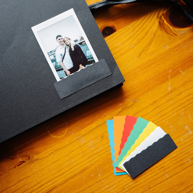 May include: A black scrapbook with a photo of a couple in a Polaroid frame. The scrapbook is open and there are colorful paper scraps on the table.