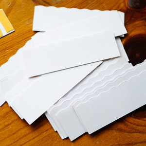 May include: A stack of white paper labels with scalloped edges. The labels are blank and ready to be used for writing or decorating.