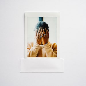 May include: A person with dark hair is wearing a light brown sweater and covering their face with their hands. They are wearing several rings on their fingers.