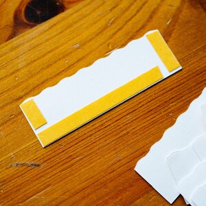 May include: White card with scalloped edges and two yellow adhesive strips. The card is on a wooden surface.