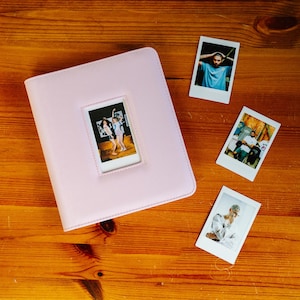 Peut inclure: Un album photo rose clair avec une fenêtre rectangulaire sur la couverture avant affichant une petite photographie de deux personnes. Trois autres petites photographies rectangulaires sont placées à droite de l'album, montrant des personnes.
