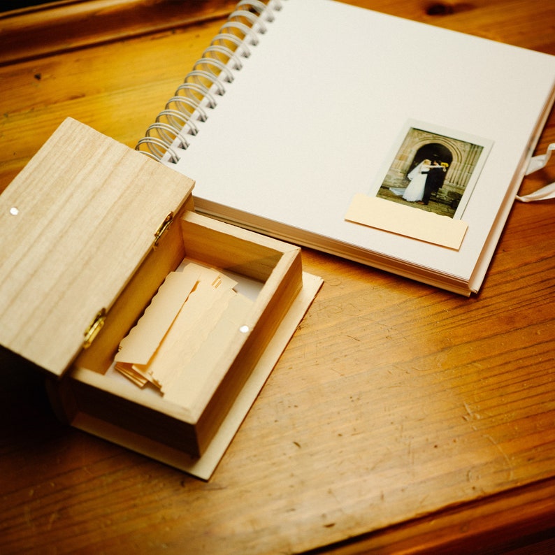 May include: A wooden box with a hinged lid, open to reveal a stack of blank cards. The box is sitting on a wooden table next to a white spiral bound notebook with a photo of a wedding couple on the cover.