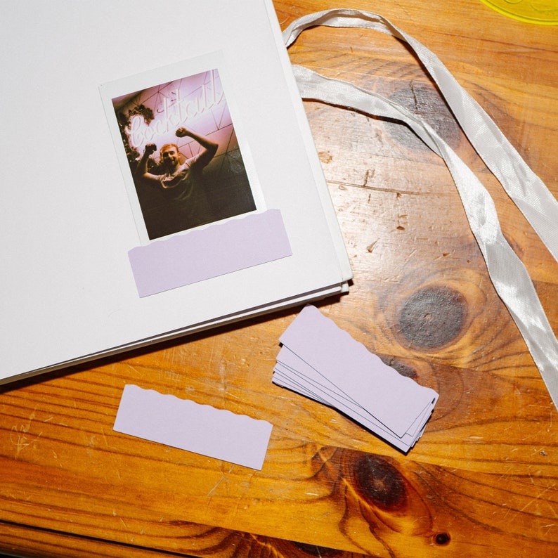 May include: A white photo album with a lavender photo corner and a photo of a person in a bar with a neon sign that says "Cocktail". The album is on a wooden surface with a white ribbon.