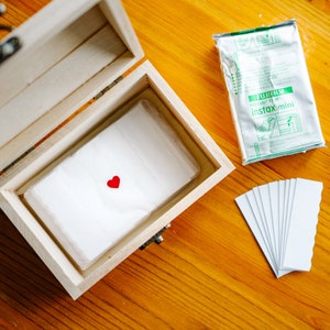 May include: A wooden treasure chest with a white tissue paper inside. The tissue paper has a red heart sticker on it. The chest is open and the lid is propped up. There is a package of Fujifilm Instax Mini instant film and a stack of white photo frames on the table next to the chest.