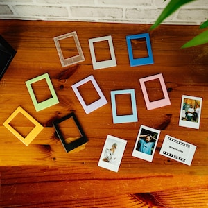 May include: A collection of rectangular plastic frames in various solid colors including yellow, green, black, white, blue, lavender, and pink, alongside three small rectangular photographs, one of which features the text RETROZI.COM.