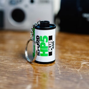 May include: A black and white film canister keychain with a green and white label that reads "Ilford HP5 Plus 400".