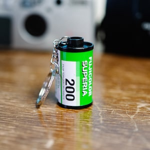 Puede incluir: Un llavero de cartucho de película verde y negro con el texto "Fujicolor Superia 200" y "Film para impresiones en color".