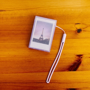 Peut inclure: Un étui photo en plastique violet avec une bordure blanche et une photo de la tour Eiffel à l'intérieur. L'étui est muni d'un cordon blanc.