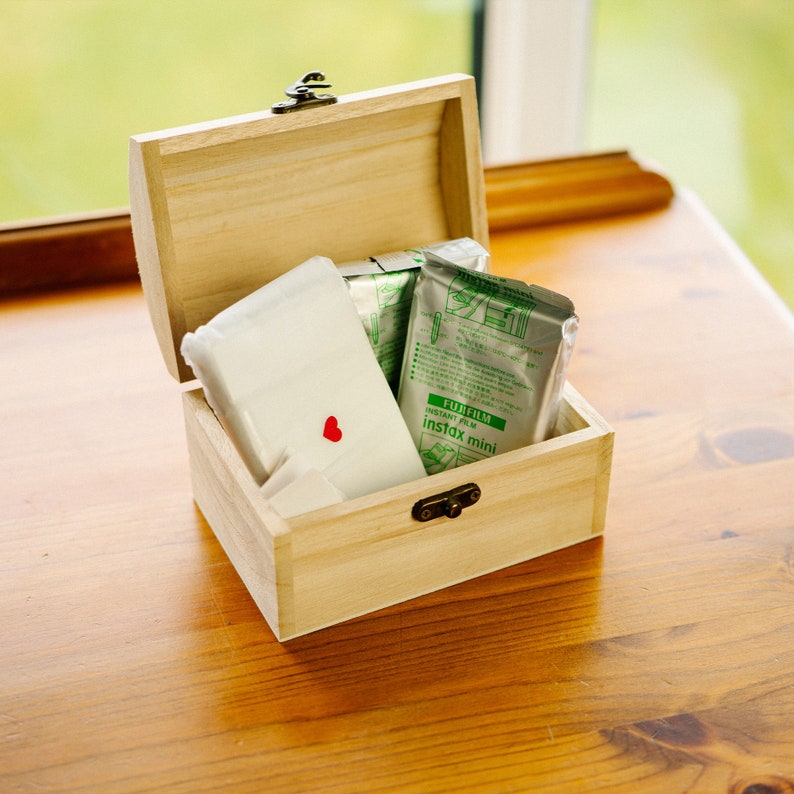 May include: A wooden box with a hinged lid, containing two packs of Fujifilm Instax Mini instant film and a white envelope with a red heart sticker.