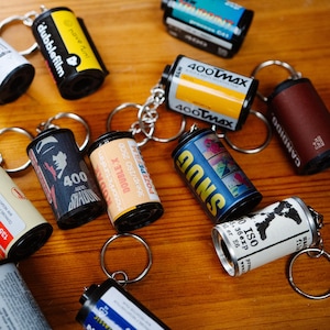 May include: A collection of vintage film canisters with keychains attached. The canisters are various colors and have different text on them, including "400TMAX", "dubblefilm", "POZEPEFILM", and "SNUG".