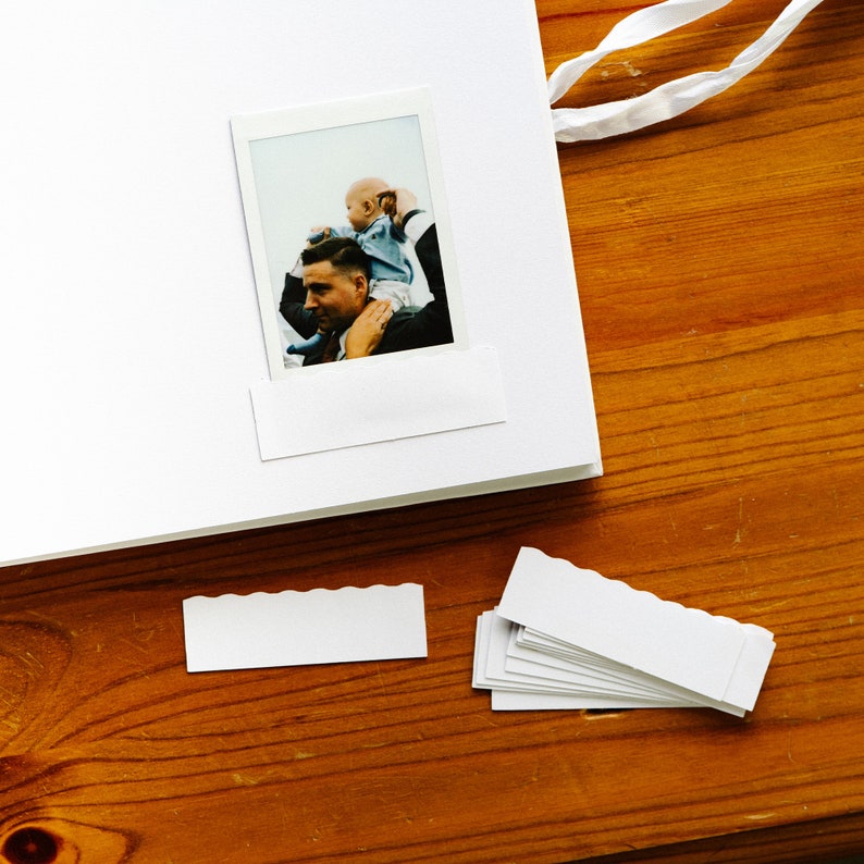 May include: A white photo album with a white photo of a man holding a baby on his shoulders. The photo is in a white frame with a white border. The album is on a wooden surface.