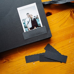 May include: A black photo album with a white photo of a couple standing together. The photo is of a man and a woman, both wearing casual clothing. The photo album is closed and has a black ribbon tied around it.