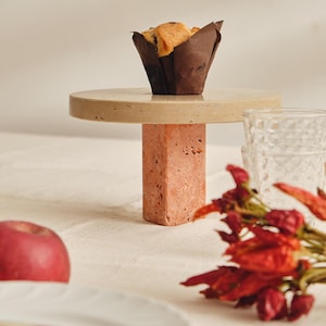 May include: A muffin in a brown paper cup sits on a small, round, beige cake stand with a square, coral-coloured base. A red apple, a glass, and red chilli peppers are also in the image. The scene is set on a white tablecloth.