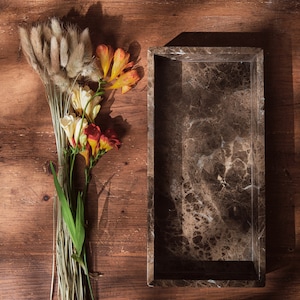 Può includere: Un vassoio rettangolare in marmo con un motivo marrone e bianco è appoggiato su una superficie di legno. Un bouquet di code di coniglio essiccate, fiori gialli, arancioni e rossi con steli verdi sono disposti a sinistra del vassoio. Il legno ha un tono caldo e naturale.