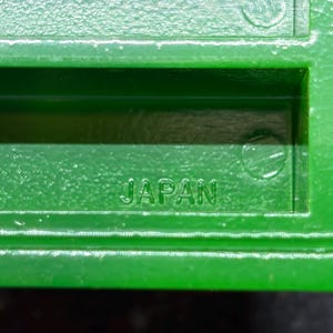 Op de afbeelding: Close-up van een heldergroen plastic object met het woord "JAPAN" in reli&euml;f. Het object heeft een rechthoekige vorm met een getextureerde afwerking en lijkt een onderdeel van een groter item te zijn.