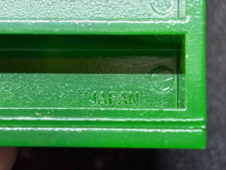 Op de afbeelding: Close-up van een heldergroen plastic object met het woord "JAPAN" in reli&euml;f. Het object heeft een rechthoekige vorm met een getextureerde afwerking en lijkt een onderdeel van een groter item te zijn.