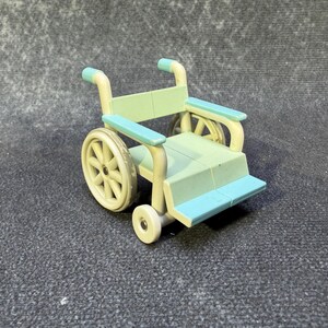 May include: A miniature wheelchair in pastel colors. The toy has a light green seat and back, with blue armrests and footrests. The wheels are a cream color, and the frame is a light yellow. The small wheelchair is a toy.