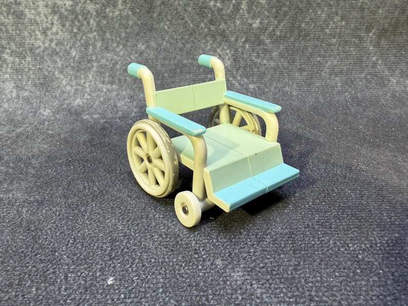 May include: A miniature wheelchair in pastel colors. The toy has a light green seat and back, with blue armrests and footrests. The wheels are a cream color, and the frame is a light yellow. The small wheelchair is a toy.