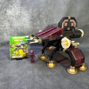 May include: A vintage Constructa toy set featuring a black and purple space vehicle with a small purple figure and a box with the text "Phantom Assault Assault Fantome". The toy has multiple parts and a detailed design.