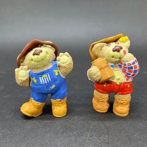 Op de afbeelding: Twee vintage berenbeeldjes. Eén beer draagt een blauwe overall en een bruine hoed. De andere beer draagt een rode korte broek en een geruit overhemd, met een pot honing. Beide beren hebben een bruine vacht en gele laarzen.