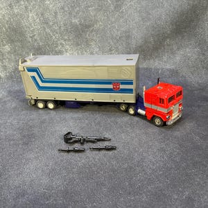 Peut inclure: Un camion semi-remorque jouet rouge et argenté avec une remorque ornée de bandes bleues et d'un insigne Autobot rouge. Le camion est accompagné de trois petits accessoires en plastique noir, dont un long fusil et deux blasters plus courts.