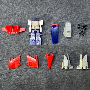 Puede incluir: Una colección de piezas de robot de juguete desmontadas en varios colores. Las piezas incluyen alas rojas, un torso azul y blanco, un escudo azul, componentes grises y un conjunto de ruedas negro. Las piezas están dispuestas sobre una superficie gris.