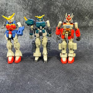 May include: Three toy robots, one blue and green, one red and white, and one red and yellow, standing on a gray surface.