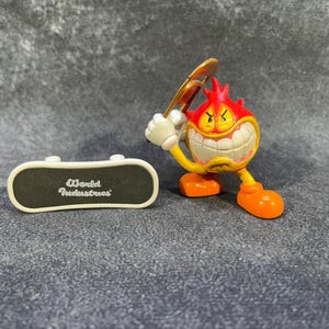 May include: A World Industries Flameboy figure with a skateboard. The Flameboy figure is red, yellow, and orange, holding a golden bat. The skateboard is black with white trim and the words "World Industries".