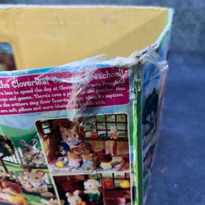 May include: A partially visible box for a Cloverleaf Corners Preschool playset. The box features illustrations of Calico Critters playing with toys and games. Text on the box reads "Welcome to the Cloverleaf Corners Preschool!"