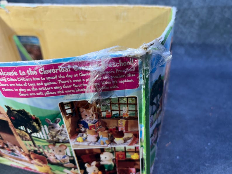 May include: A partially visible box for a Cloverleaf Corners Preschool playset. The box features illustrations of Calico Critters playing with toys and games. Text on the box reads "Welcome to the Cloverleaf Corners Preschool!"