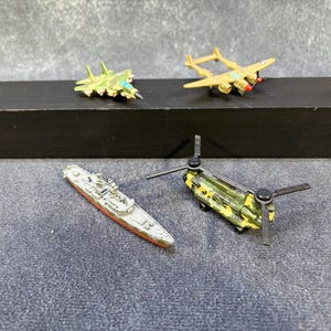 以下が含まれることがあります： 緑色の迷彩柄戦闘機、タンカラーの双胴プロペラ機、赤い船体の灰色軍艦、緑色の迷彩柄タンデムローター輸送ヘリコプターの4つのミニチュアダイキャスト製乗り物コレクション。
