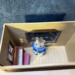 May include: A miniature dollhouse scene featuring a small, tan-colored cat figurine wearing a blue and white outfit. The scene includes a piano, a window, and a barred window. The dollhouse is beige.