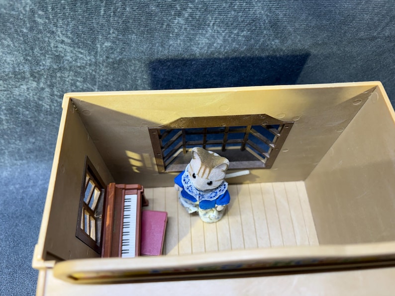 May include: A miniature dollhouse scene featuring a small, tan-colored cat figurine wearing a blue and white outfit. The scene includes a piano, a window, and a barred window. The dollhouse is beige.
